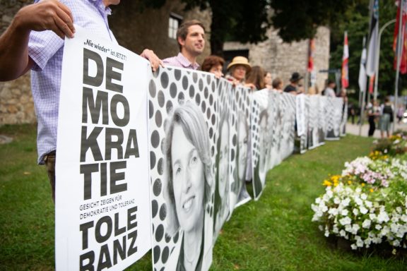 Menschen halten lange Reihe von Plakaten mit Porträts und Schriftzug ‚Demokratie Toleranz‘ bei Freiluftveranstaltung.