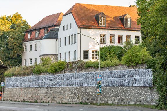 An der Stadtmauer hängt langes Banner mit vielen schwarz-weißen Porträts unter weißem Gebäude mit rotem Dach.