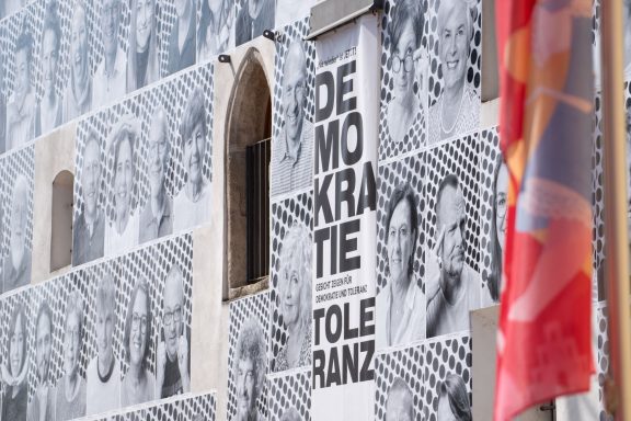 Fassade des Herzogskastens in Abensberg mit großflächigen Schwarz-weiß-Porträts und Schriftzug ‚DEMOKRATIE TOLERANZ‘.