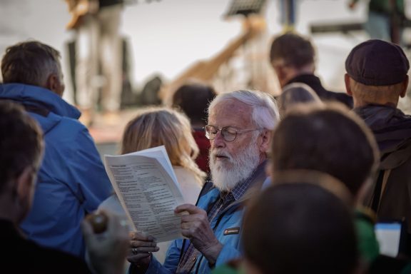Gruppe von Menschen von hinten und seitlich, eine Person hält gefaltetes Papier mit Liedtexten.