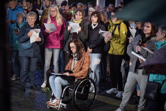 Gruppe von Menschen singt mit Liedtexten in der Hand, eine Person sitzt im Rollstuhl mit Liedblatt.