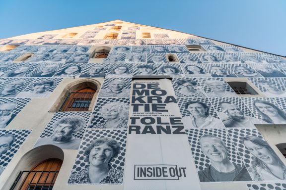 Fassadeninstallation mit vielen schwarz-weißen Porträts, Schriftzug ‚DEMOKRATIE TOLERANZ‘ und ‚INSIDE OUT‘.