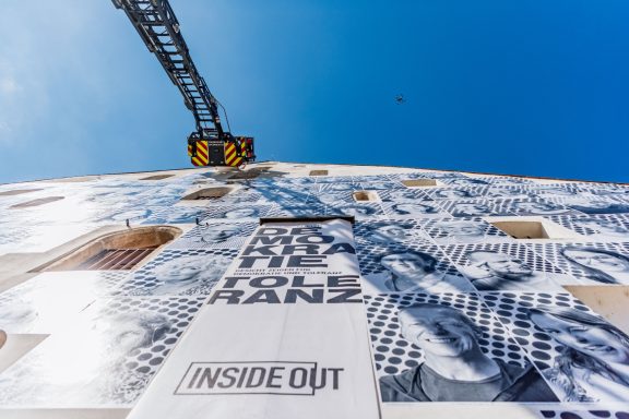 Blick von unten auf hohe Fassade mit Schwarz-weiß-Porträts und Schriftzug ‚DEMOKRATIE TOLERANZ‘, darüber Feuerwehrleiter.