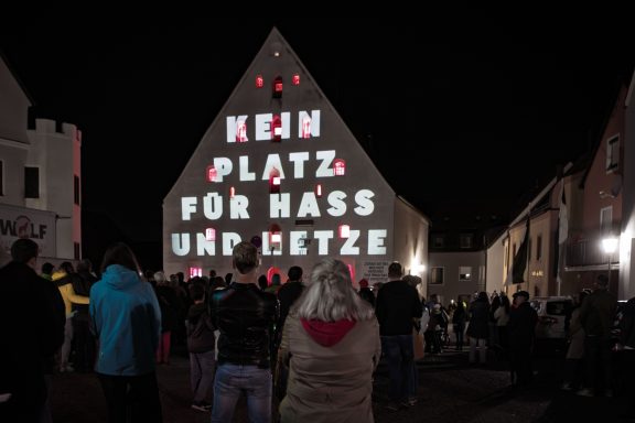 Fassade des Herzogskastens mit Lichtinstallation und Botschaft ‚KEIN PLATZ FÜR HASS UND HETZE‘ vor Menschenmenge bei Nacht.