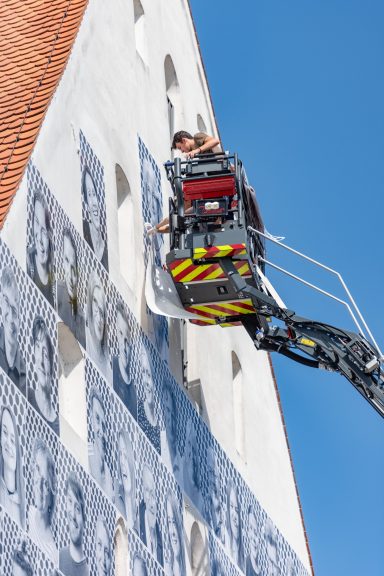 Person auf Drehleiter bringt großformatige Schwarz-weiß-Porträts an der Fassade des Herzogskastens an.