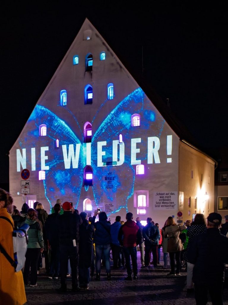 Menschenmenge vor dem Herzogskasten mit Laserprojektion ‚NIE WIEDER!‘ auf der Fassade bei Nacht.
