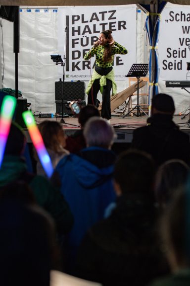 RiA REiSER steht auf Bühne vor Mikrofon, trägt gemustertes Oberteil und helles Kleidungsstück um die Hüfte.