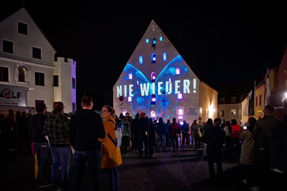 Fassade des Herzogskastens mit Lichtinstallation und Botschaft ‚NIE WIEDER‘ vor Menschenmenge bei Nacht.