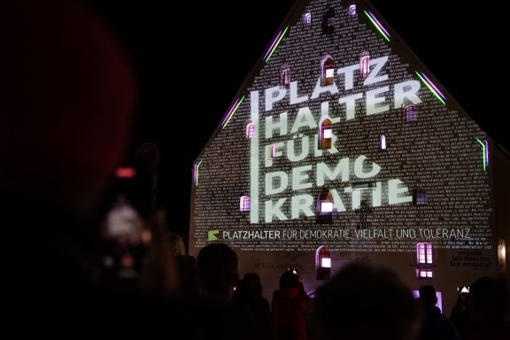 Licht- und Laserinstallation mit Schriftzug ‚PLATZHALTER FÜR DEMOKRATIE, VIELFALT UND TOLERANZ‘, davor Menschenmenge.