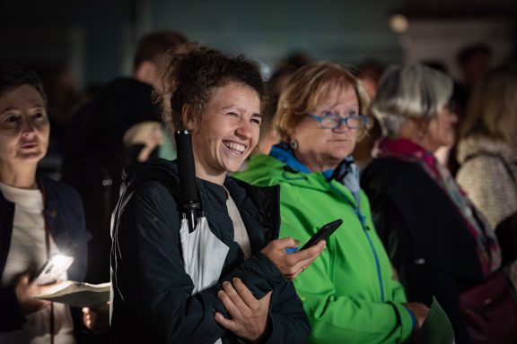 Mehrere Personen stehen eng beieinander, eine hält Smartphone und Regenschirm, alle blicken nach vorne.