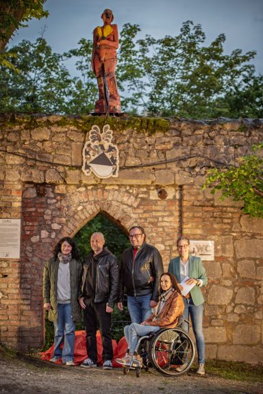 Fünf Personen vor alter Steinmauer mit Wappen und Statue, eine Person sitzt im Rollstuhl.