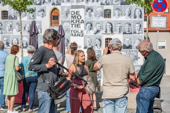 Gruppe vor Fassade mit Schriftzug ‚DEMOKRATIE TOLERANZ‘, darunter Filmcrew mit Mikrofon und Kamera bei Straßeninterview.