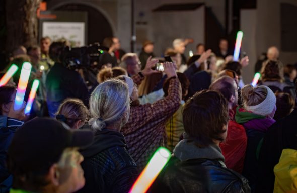 Gruppe von Teilnehmern am PopUp Chor bei Nacht mit leuchtenden Stäben in der Hand und erhobenen Armen