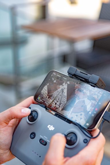 Hände steuern DJI-Drohnenfernbedienung mit Smartphone, das Luftaufnahme des Herzogskastens zeigt.