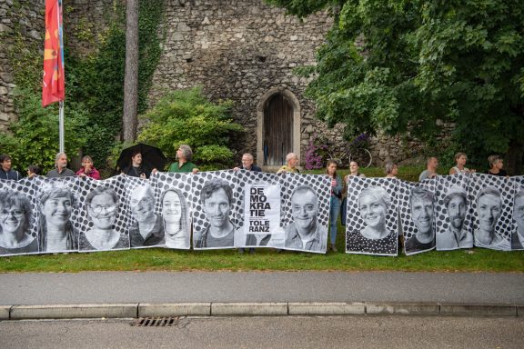 Menschenkette vor Steinmauer mit Tür hält langes Banner mit vielen schwarz-weißen Porträts und Schriftzug ‚DEMOKRATIE‘.