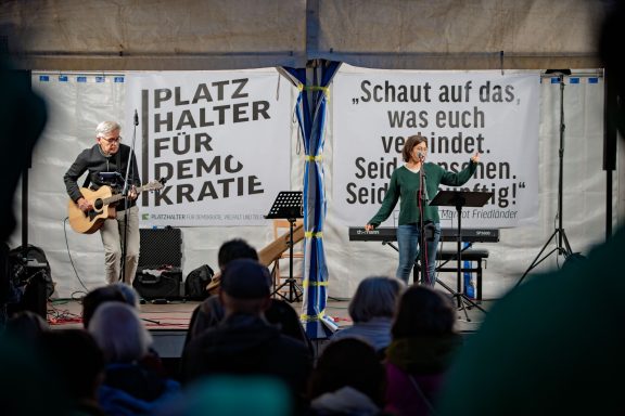 Zwei Personen auf Bühne vor Bannern, eine spielt Gitarre, die andere spricht ins Mikrofon, Publikum im Vordergrund.