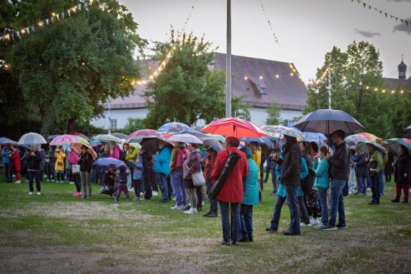 Gruppe von Menschen mit Regenschirmen steht auf Wiese vor Gebäude mit Lichterketten im Hintergrund.