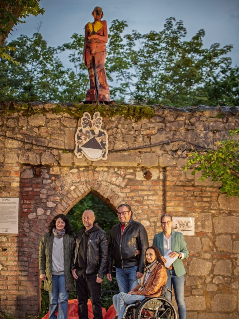 Fünf Personen vor alter Steinmauer mit Wappen und Holzskulptur ‚Davina‘ auf der Schlossgartenmauer.