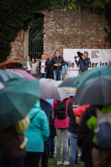 Menschen mit Regenschirmen stehen vor fünf Personen vor alter Steinmauer mit Banner im Hintergrund.