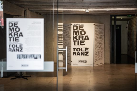 Großes Ausstellungsplakat mit den Worten ‚DEMOKRATIE‘ und ‚TOLERANZ‘ im Museum, dazu Texte und Zitate zu Demokratie.
