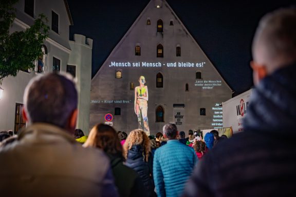 Fassade des Herzogskastens mit Licht- und Laserinstallation, zeigt Skulptur ‚DAVINA‘ und Text ‚Mensch sei Mensch – und bleib es‘.