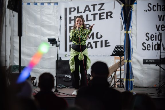 Person mit gemustertem Oberteil steht auf Bühne vor Banner ‚PLATZ HALTER DEMO KRATIE‘ und singt ins Mikrofon.